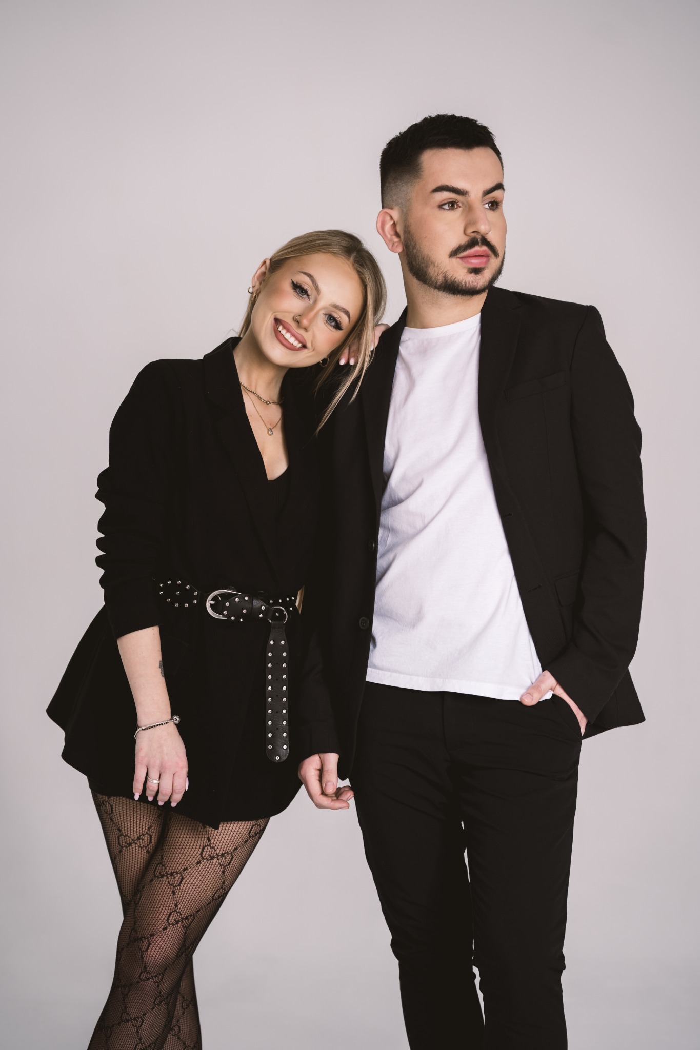 Portret eleganckiej pary: blondynka w czarnej marynarce z ozdobnym paskiem i rajstopach w logo, oparta o ramię bruneta w białym t-shircie i czarnej marynarce, na jasnym tle.