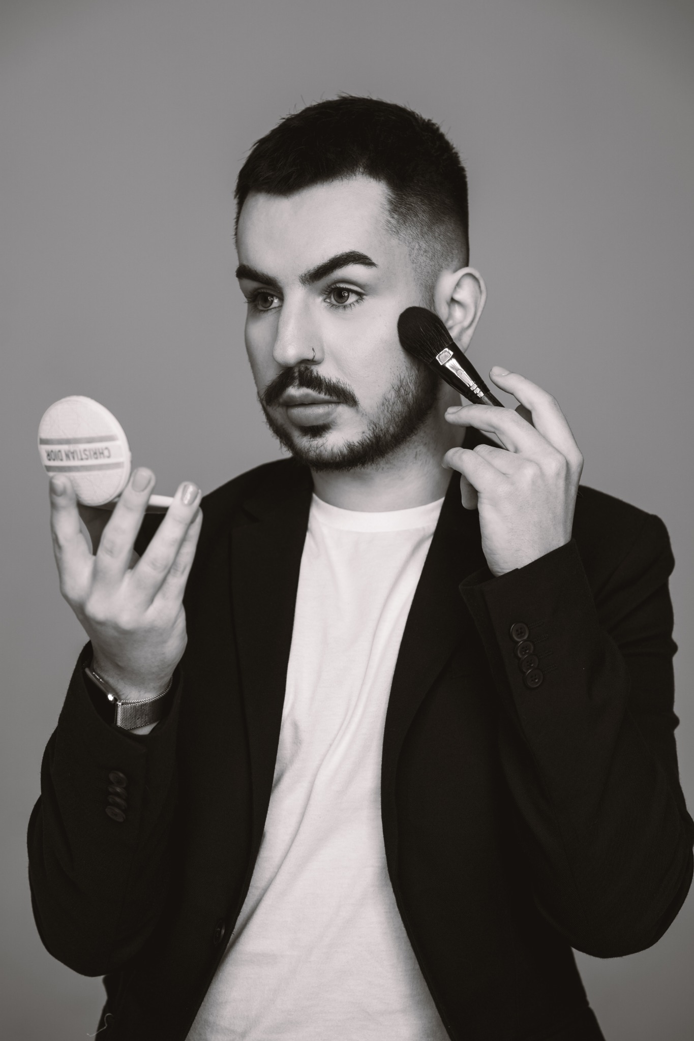 Czarno-białe zdjęcie mężczyzny w marynarce, który wykonuje makijaż pędzlem, patrząc w lusterko z napisem CHRISTIAN DIOR.