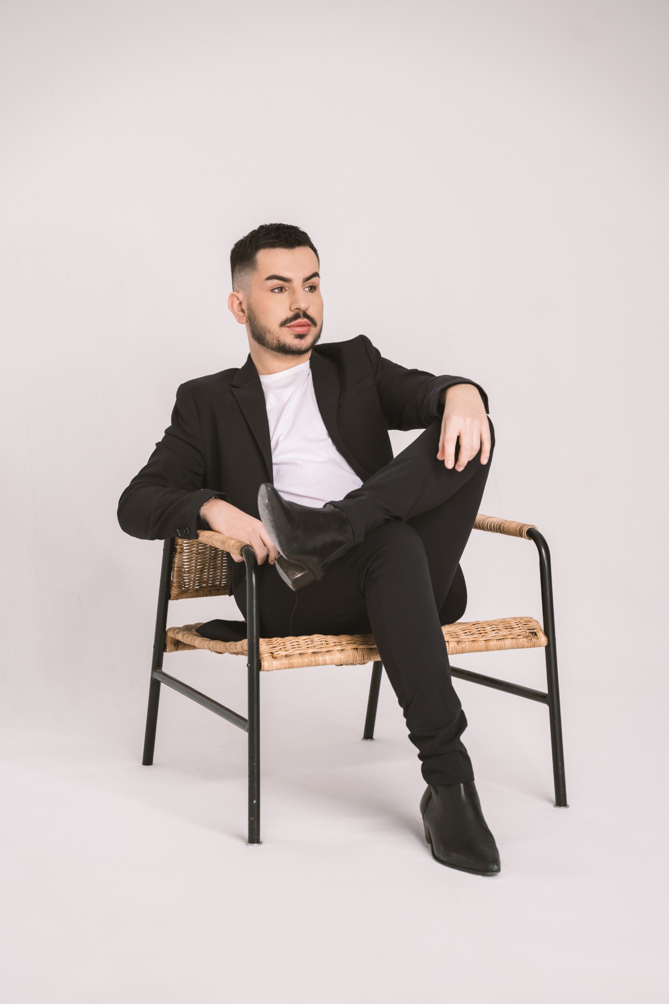 Mężczyzna z brodą, ubrany w czarny garnitur i biały t-shirt, siedzi na wiklinowym krześle na białym tle, z nogami skrzyżowanymi w kostkach i wzrokiem skierowanym w bok.