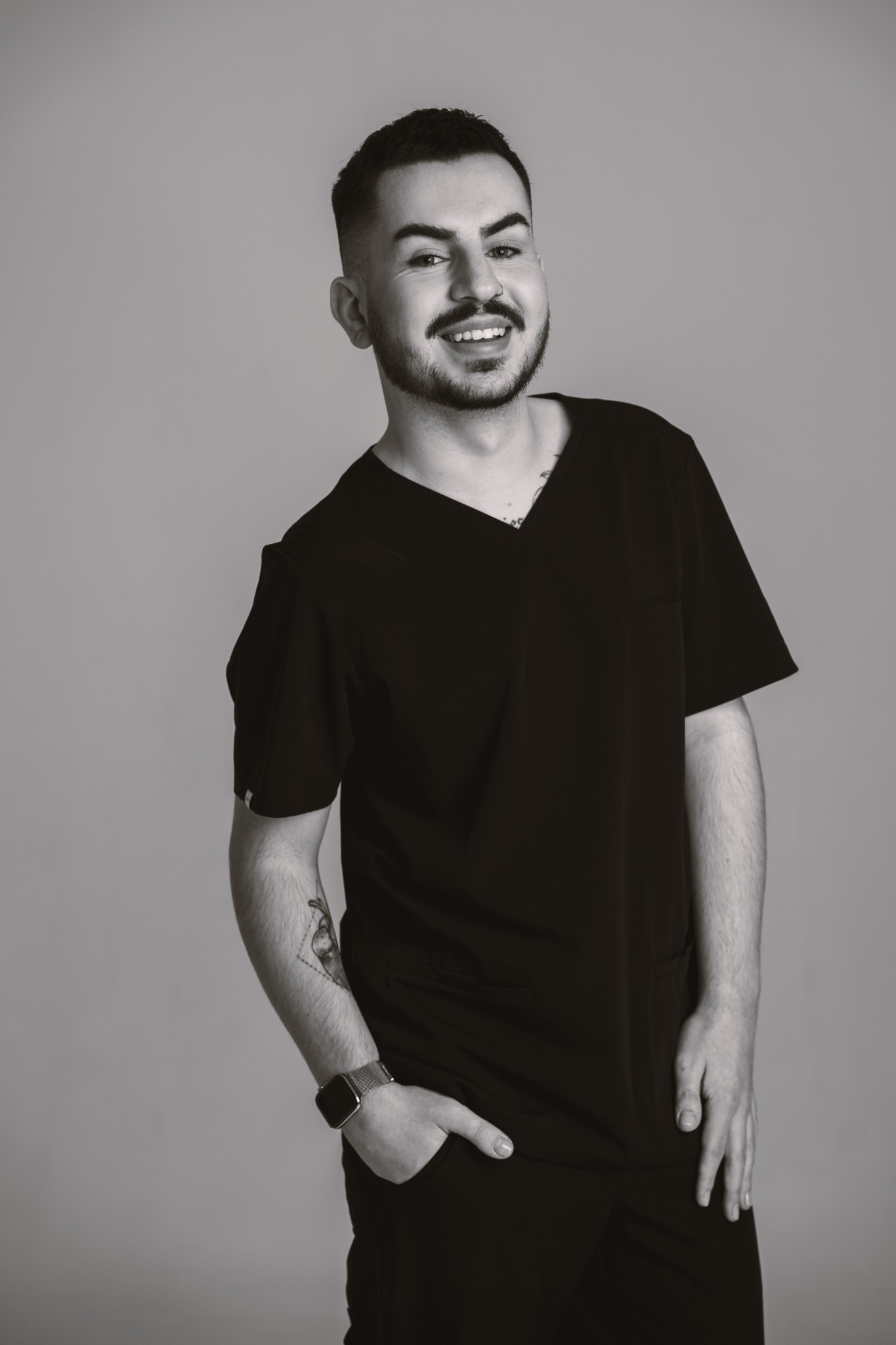 Portret uśmiechniętego mężczyzny w czarnym uniformie medycznym, z tatuażem na przedramieniu i zegarkiem na ręce, na jednolitym szarym tle. Zdjęcie czarno-białe.