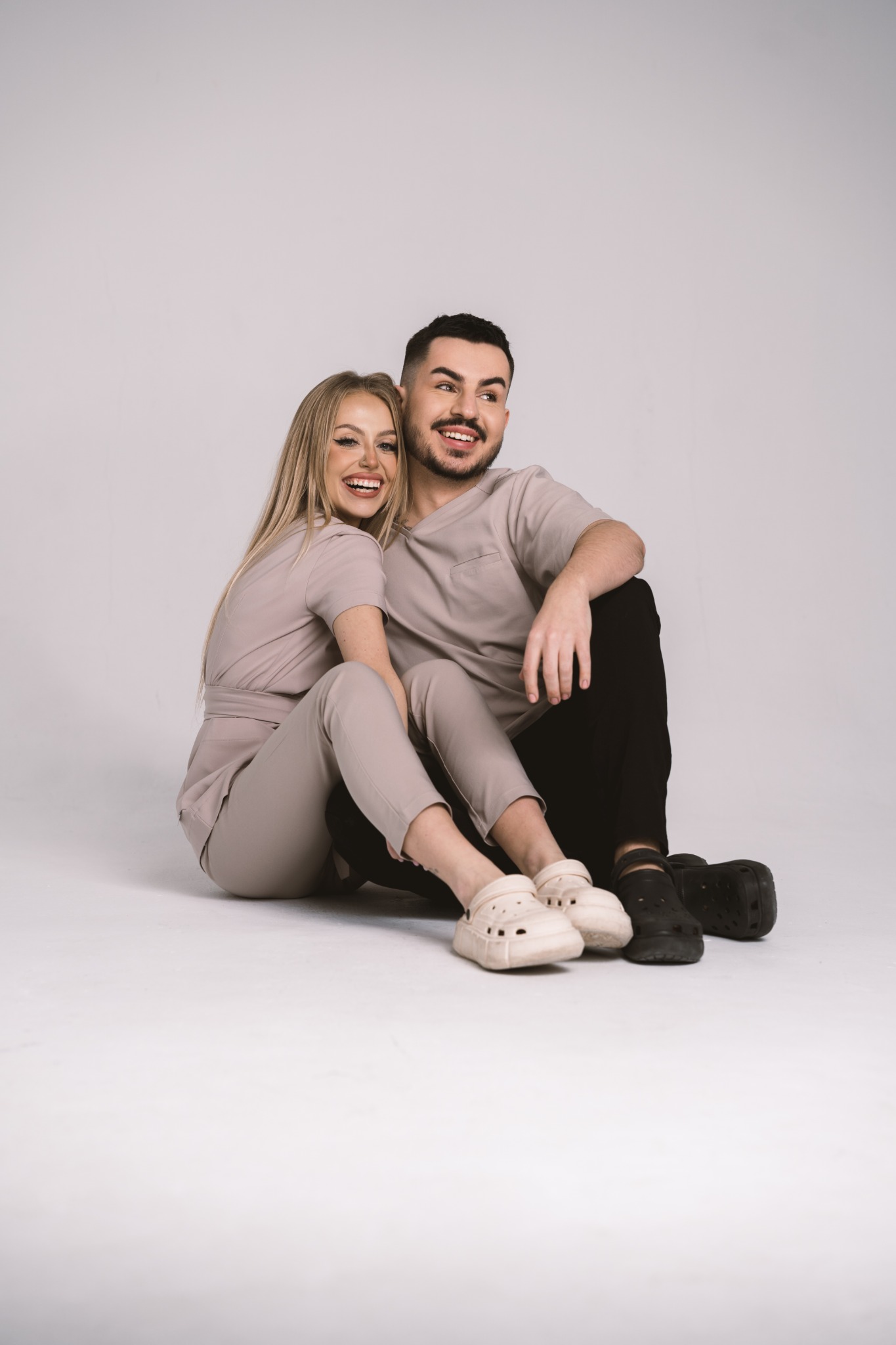Uśmiechnięta blondynka i brunet siedzą na podłodze w dopasowanych beżowych uniformach medycznych i obuwiu typu Crocs, pozują na jasnym tle.