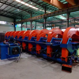 Maszyna do gięcia blachy w hali produkcyjnej, pomalowana na niebiesko i pomarańczowo, z widocznym systemem hydraulicznym i wentylatorem chłodzącym, na tle metalowej konstrukcji dachu.