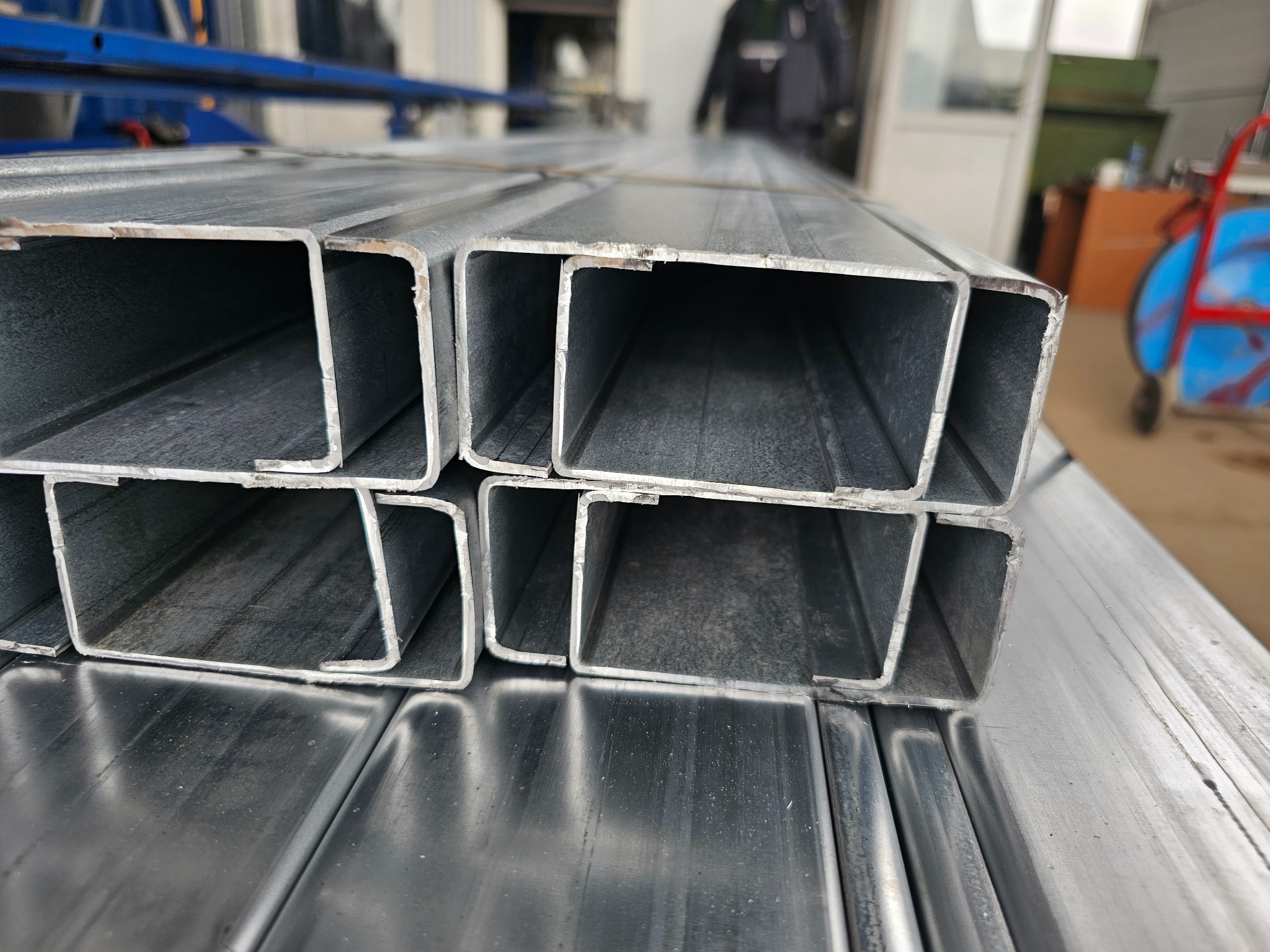 Stos ocynkowanej blachy trapezowej o profilu C, widoczny z bliska, zarysowana powierzchnia, w tle warsztat z wózkiem transportowym.