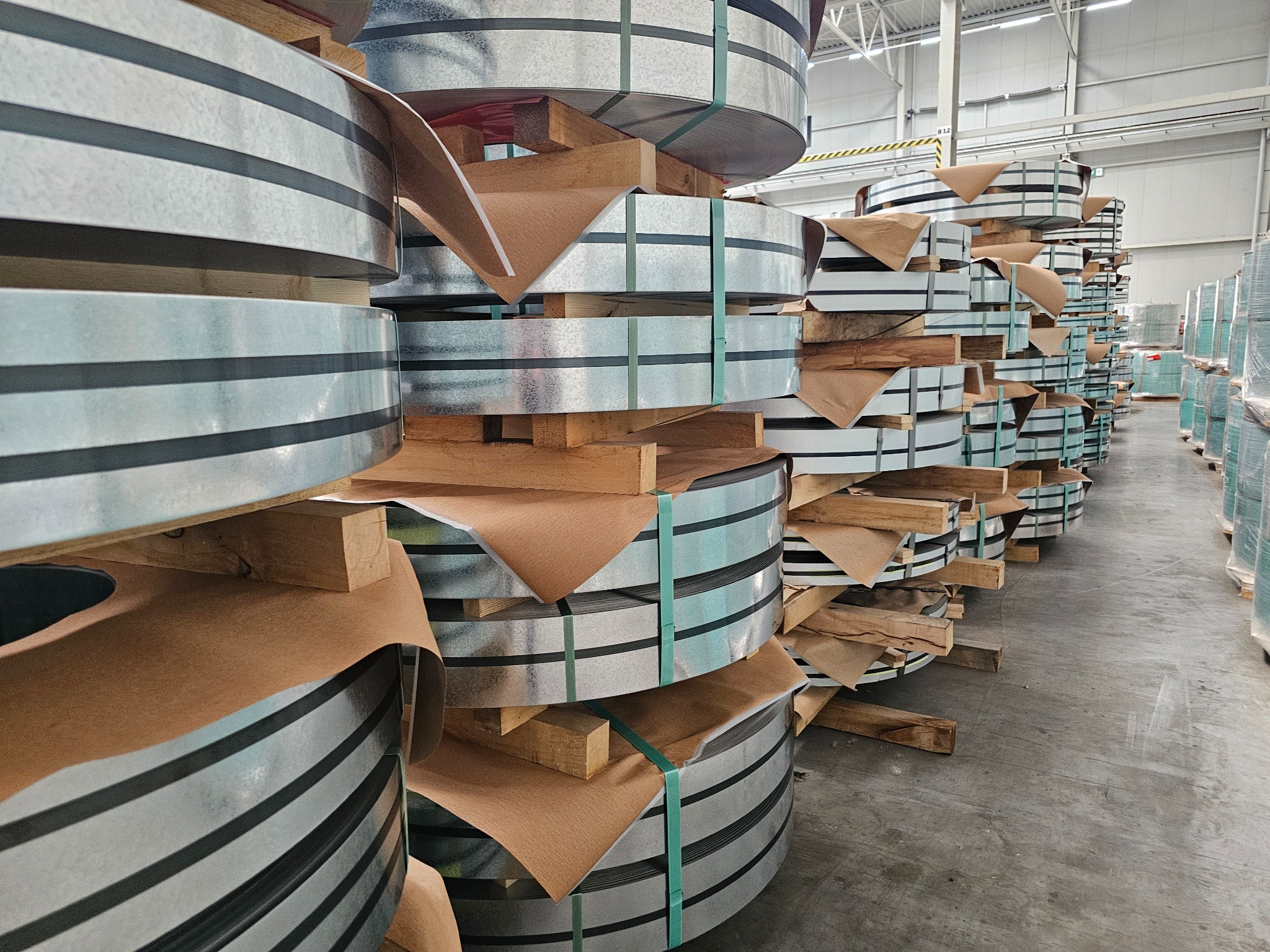 Stosy kręgów blachy ocynkowanej zabezpieczonych brązowym papierem i spiętych zieloną taśmą, ułożone na drewnianych paletach w magazynie.