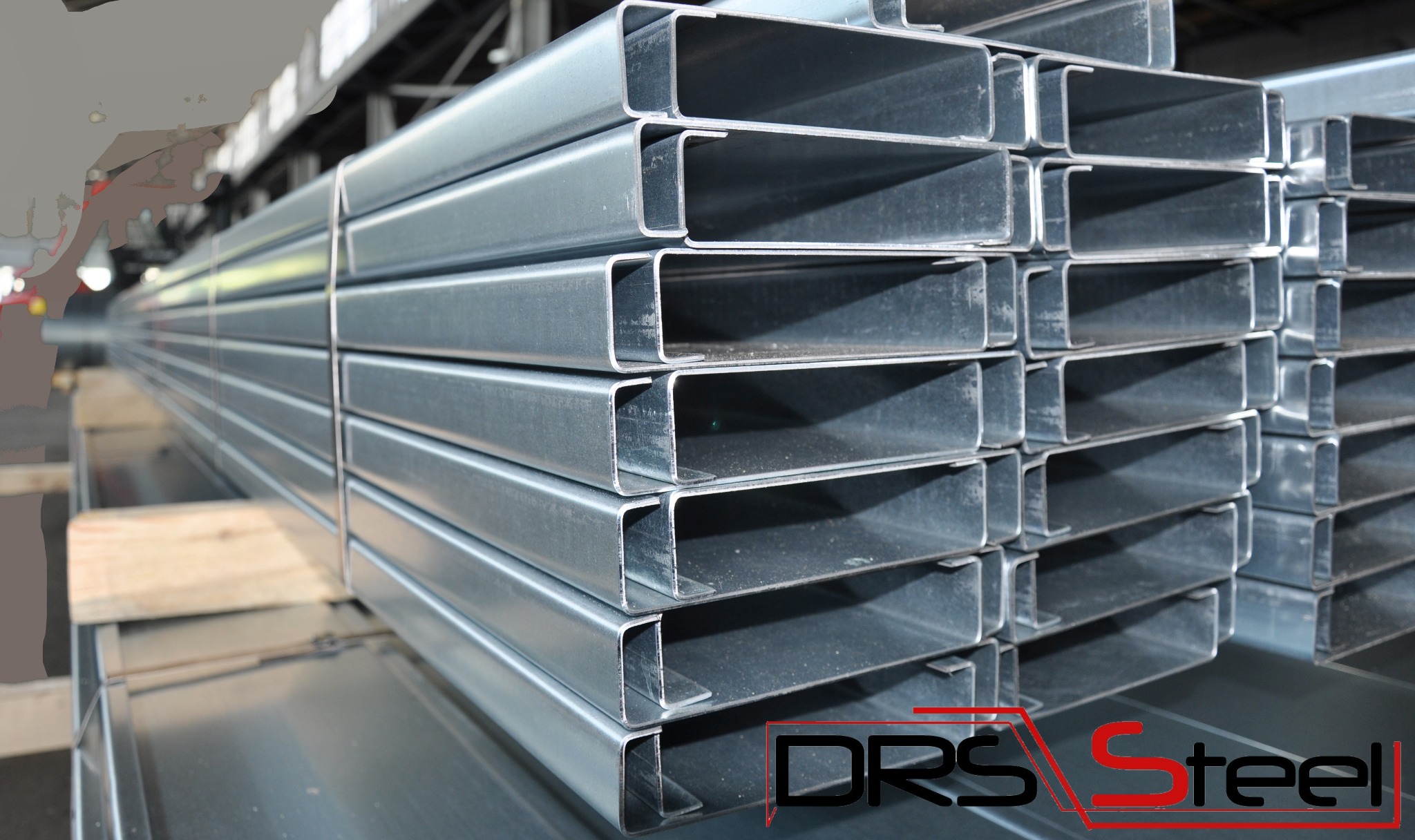 Stos ocynkowanych profili stalowych o przekroju trapezowym, ułożonych na drewnianych legarach, z widocznym logo producenta DRS Steel w prawym dolnym rogu.