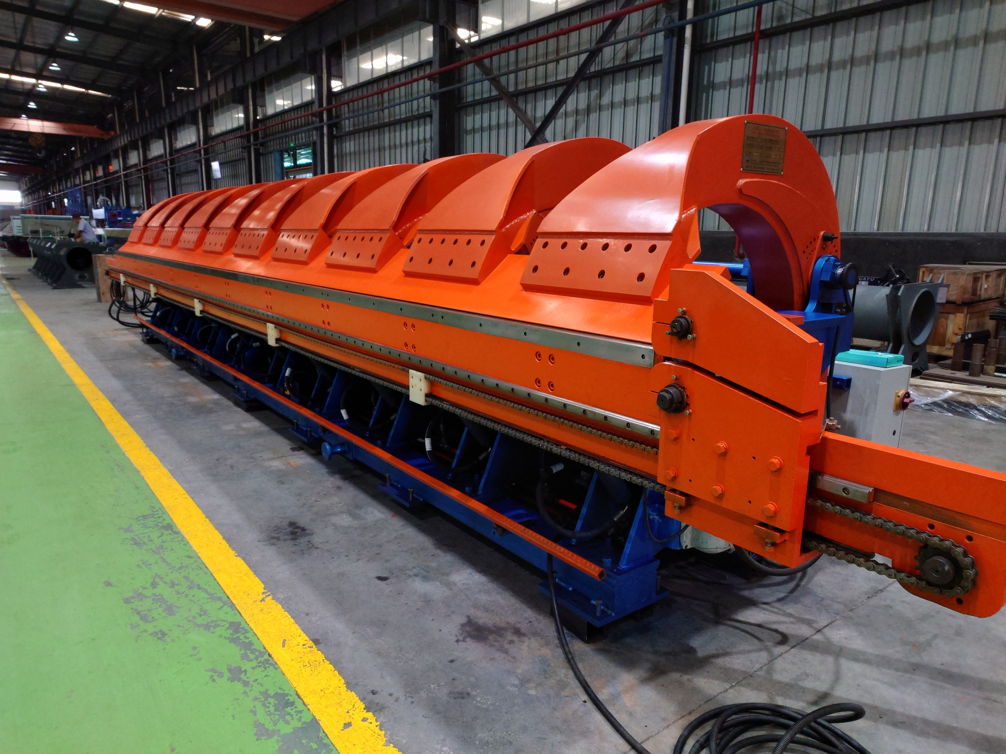 Pomarańczowa maszyna do formowania blachy trapezowej z łańcuchem napędowym, widoczna w hali produkcyjnej z zielono-szarą podłogą i żółtymi pasami.