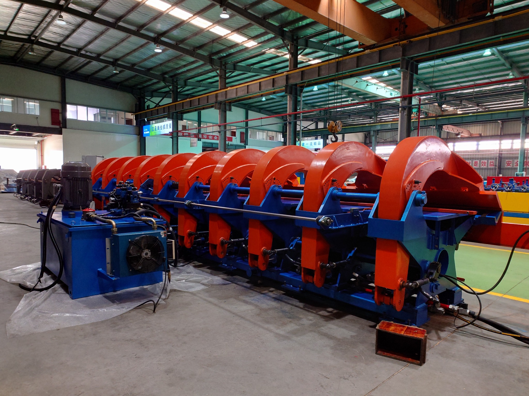 Maszyna do gięcia blachy w hali produkcyjnej, pomalowana na niebiesko i pomarańczowo, z widocznym systemem hydraulicznym i wentylatorem chłodzącym, na tle metalowej konstrukcji dachu.