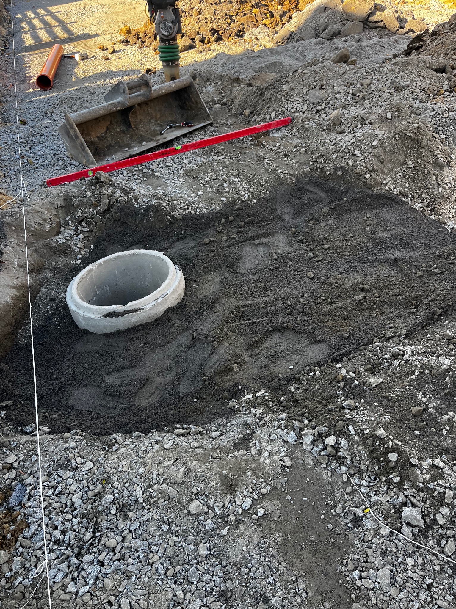 Betonowy krąg studzienny umieszczony w wykopie przygotowanym pod montaż, obok leży poziomica, a w tle widoczny sprzęt budowlany.