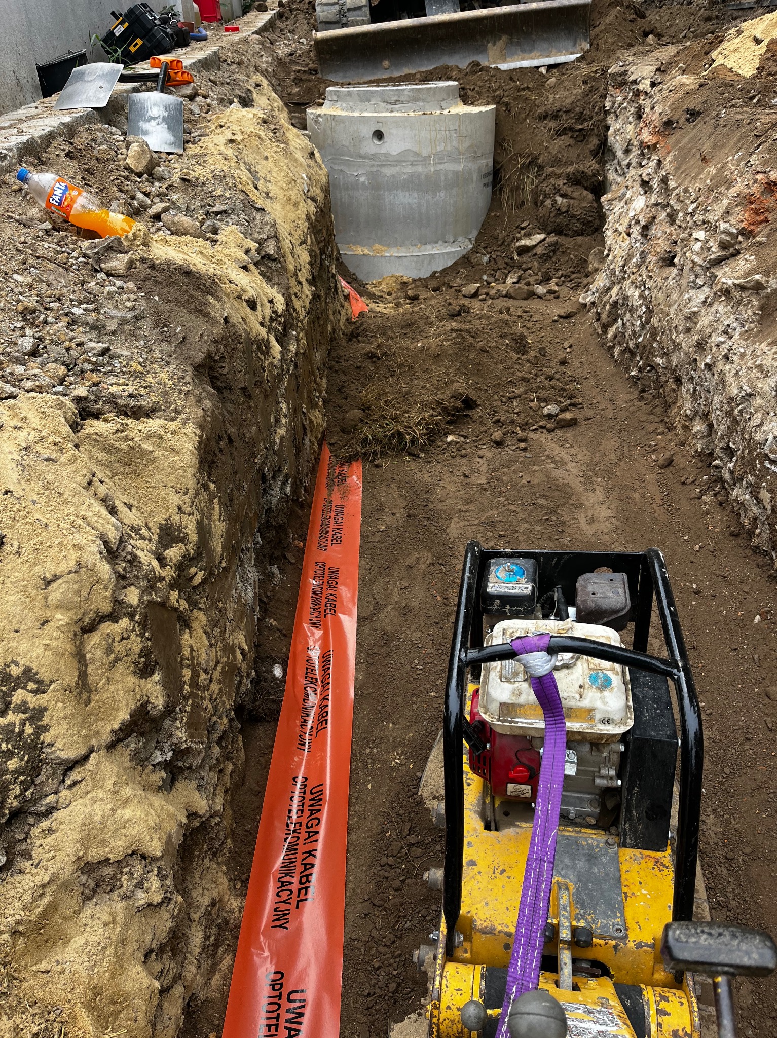 Wykonany wykop pod instalacje sanitarne z widocznym betonowym zbiornikiem, pomarańczową taśmą ostrzegawczą o kablu optotelekomunikacyjnym oraz ubijarką wibracyjną do zagęszczania gruntu.