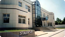 Budynek biurowy o jasnej elewacji z dużymi oknami i przeszklonym wejściem, widoczny trawnik z logo firmy STALEXPORT.
