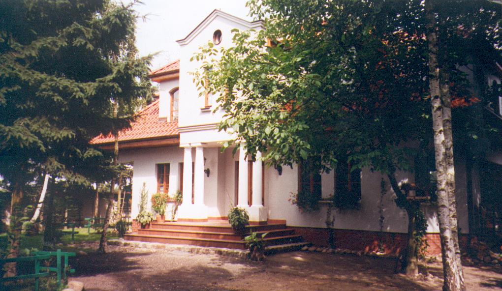 Gotowy dom murowany z kolumnami przy wejściu, czerwonym dachem i roślinami w donicach na schodach, otoczony zielenią.