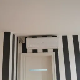 Biała klimatyzacja ANDE zamontowana nad drzwiami w pokoju z czarno-białymi pasami na ścianie, widoczny również anemostat.