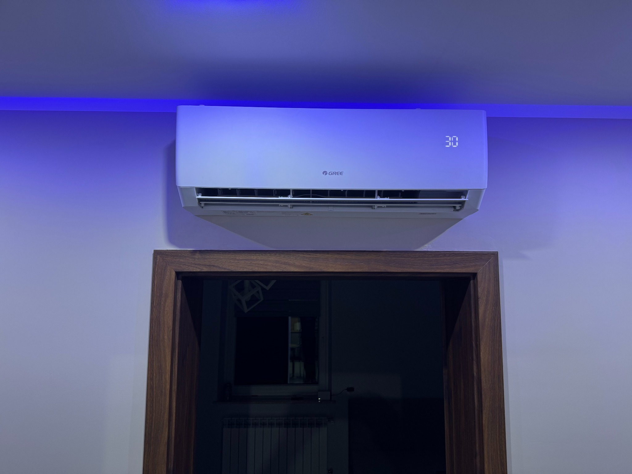 Biała klimatyzacja marki Gree zamontowana nad drewnianą framugą drzwi, podświetlona niebieskim światłem LED na suficie.