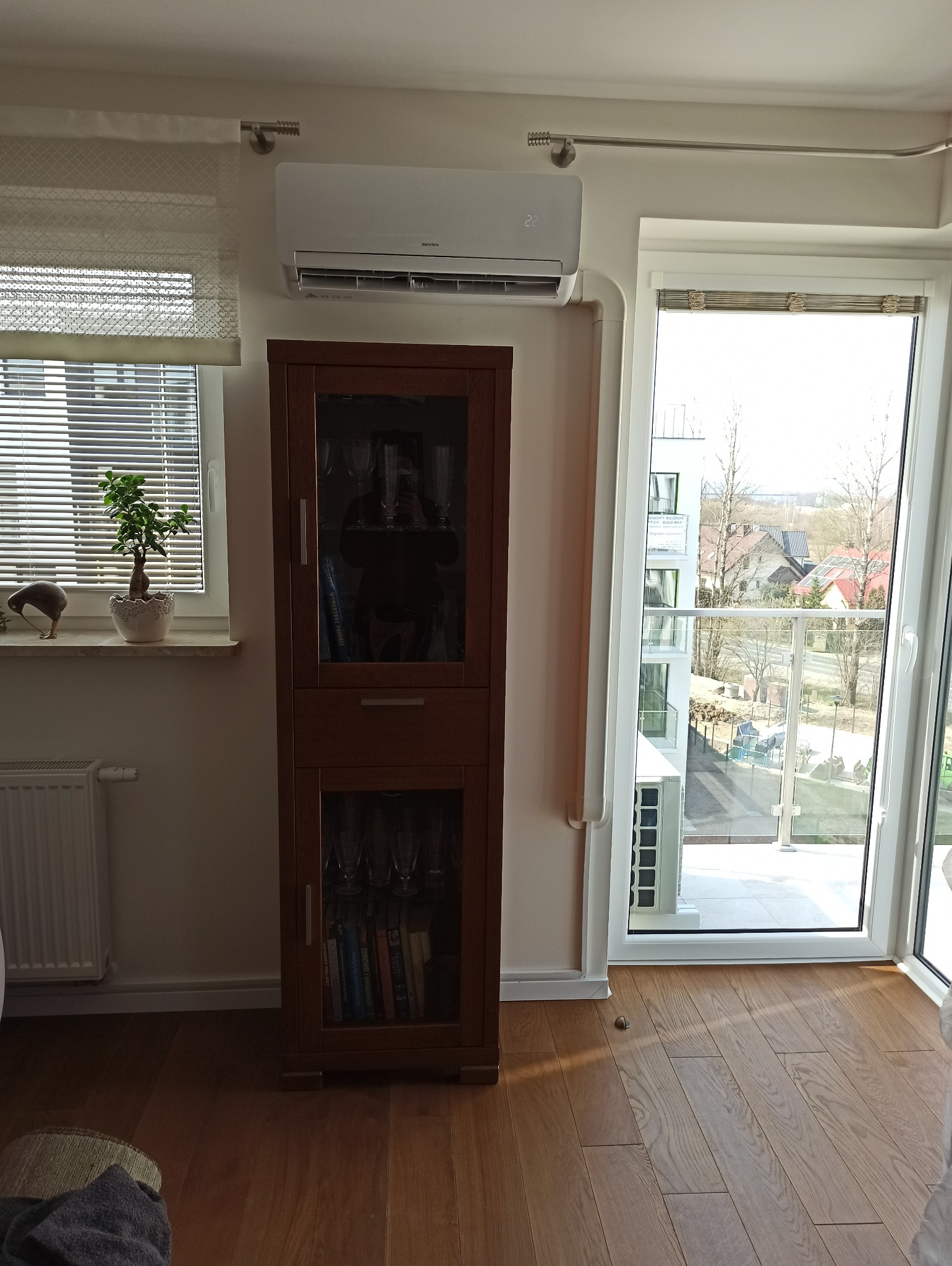Biały klimatyzator ścienny zamontowany nad drewnianą witryną w jasnym pokoju z balkonem i rośliną w doniczce na parapecie.
