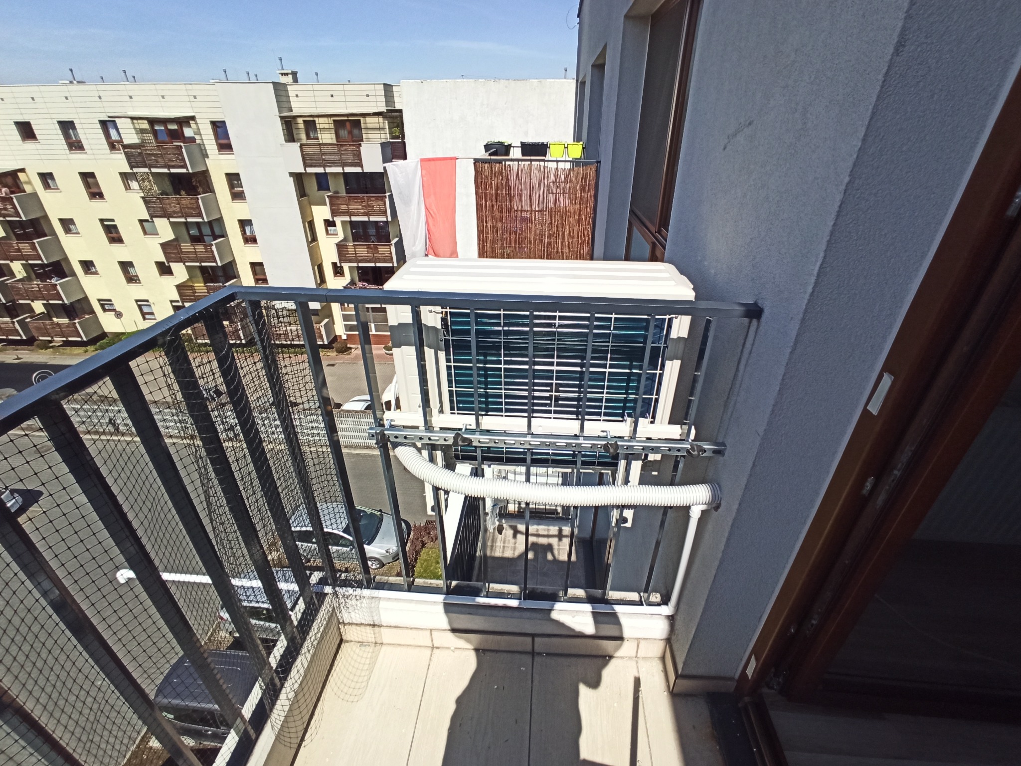 Zewnętrzna jednostka klimatyzacji zamontowana na balkonie mieszkania w bloku, widok z góry na ulicę i sąsiedni budynek.