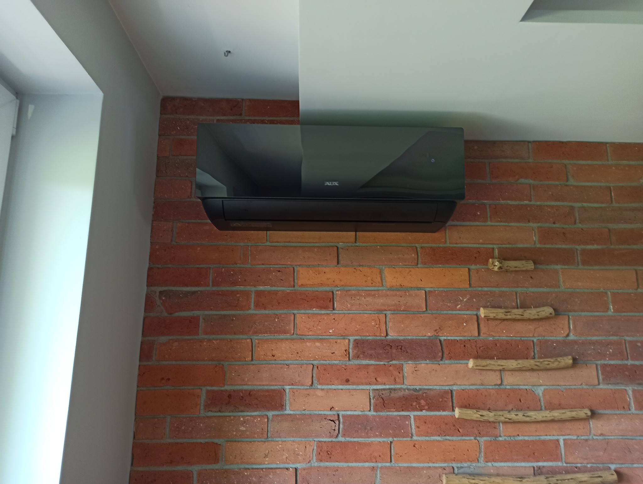 Czarna jednostka klimatyzacyjna marki AUX zamontowana na ścianie z czerwonej cegły z dekoracyjnymi drewnianymi elementami.