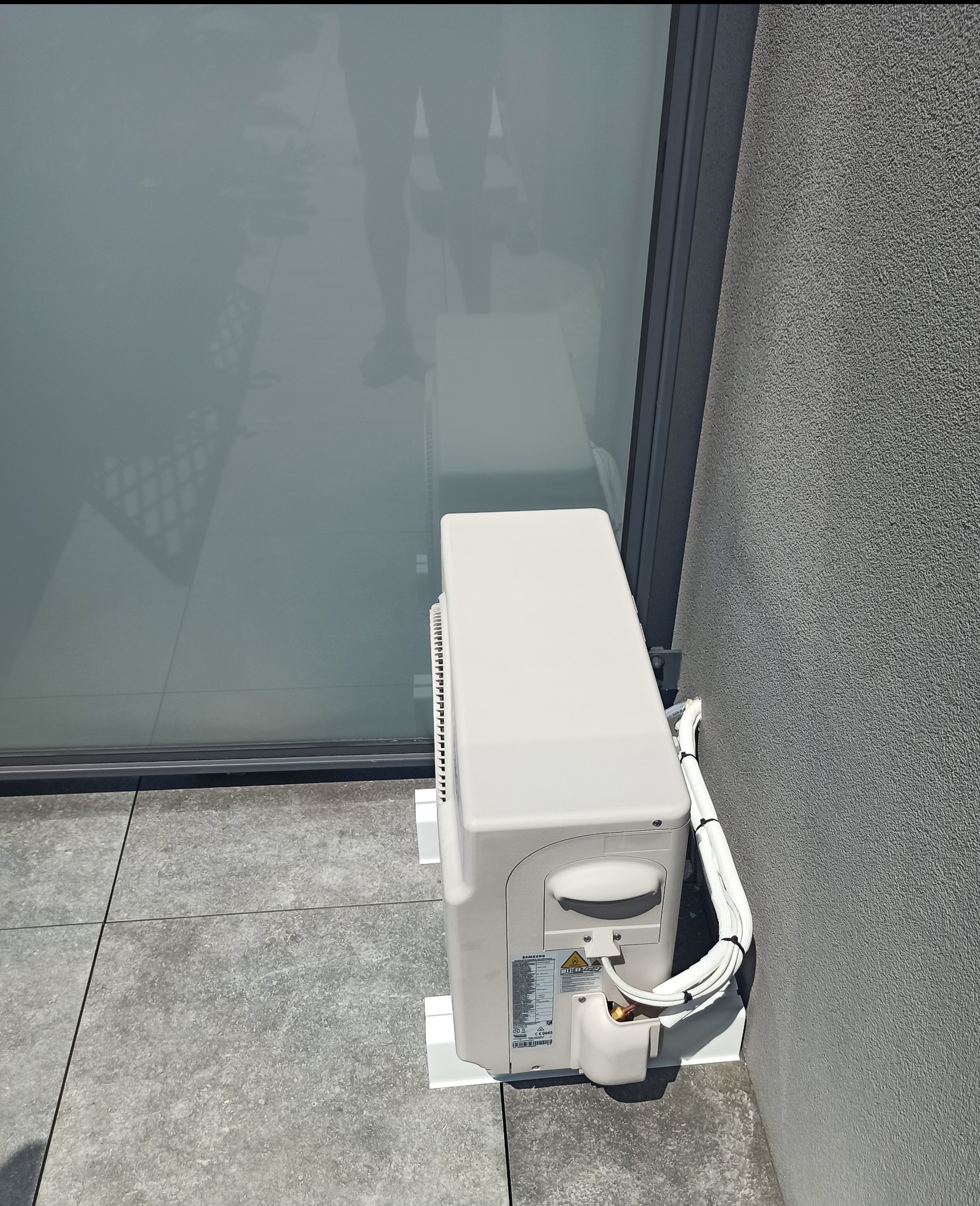 Zewnętrzna jednostka klimatyzatora Samsung zamontowana na balkonie z szarymi płytkami i widocznymi przewodami w białej osłonie przy szarej ścianie budynku.