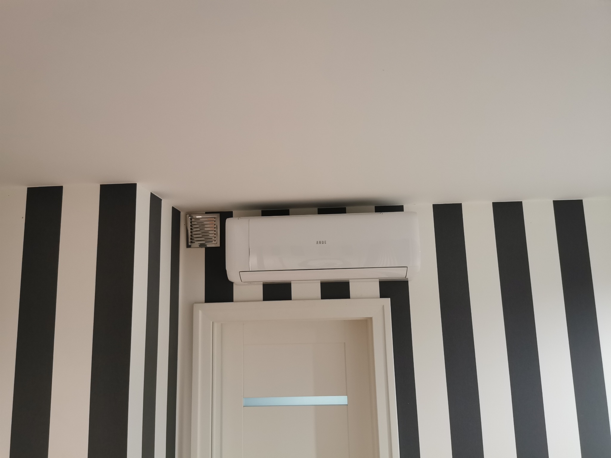 Biała klimatyzacja ANDE zamontowana nad drzwiami w pokoju z czarno-białymi pasami na ścianie, widoczny również anemostat.