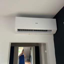 MICHAŁ SROKA - Biała klimatyzacja Haier, zamontowana na ścianie w jasnym wnętrzu. W lustrze odbija się osoba robiąca zdjęcie. Kontrastująca czarna ściana w tle.