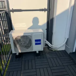 Biała jednostka zewnętrzna klimatyzacji Haier, zamontowana na balkonie z czarną podłogą, widok z boku, z widocznymi przewodami i cieniem osoby na ścianie.