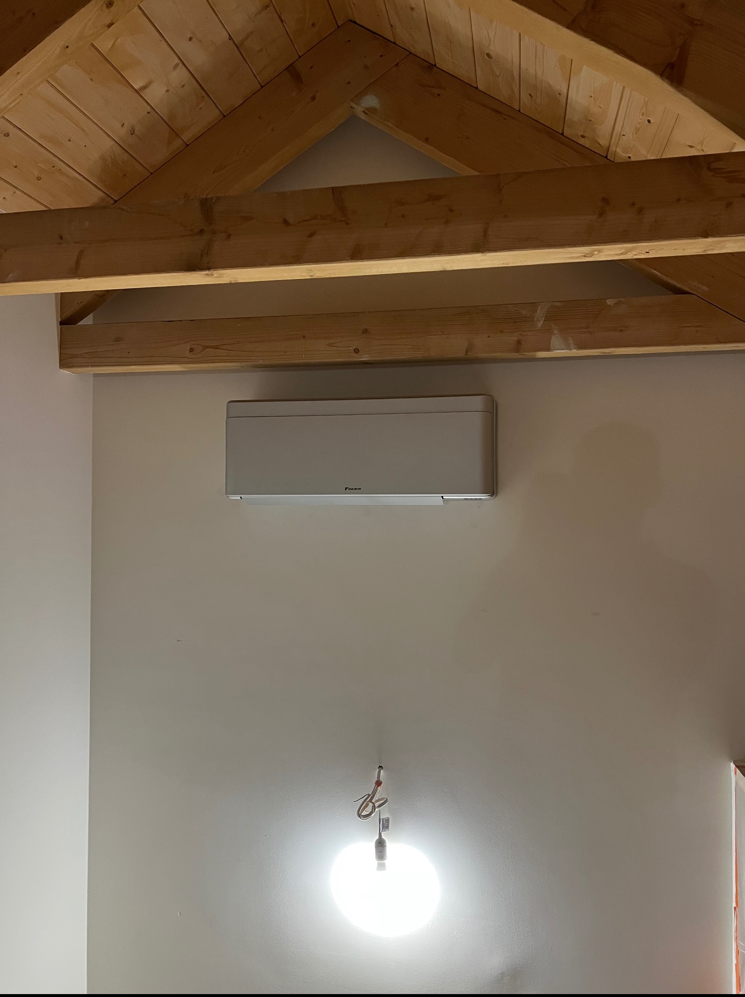 Zamontowany klimatyzator Daikin na ścianie pod drewnianą konstrukcją dachu, widoczne przewody oświetleniowe.