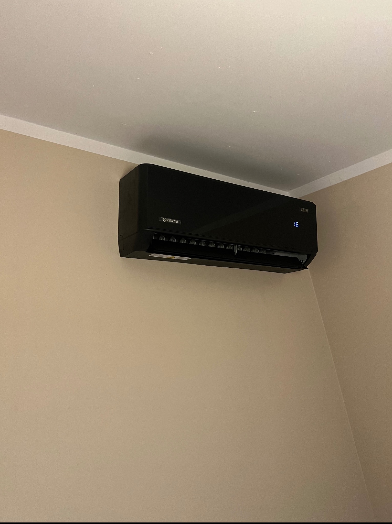 Czarna klimatyzacja Rotenso/Tesla zamontowana na beżowej ścianie w rogu pomieszczenia. Widoczny wyświetlacz z temperaturą 16 stopni.