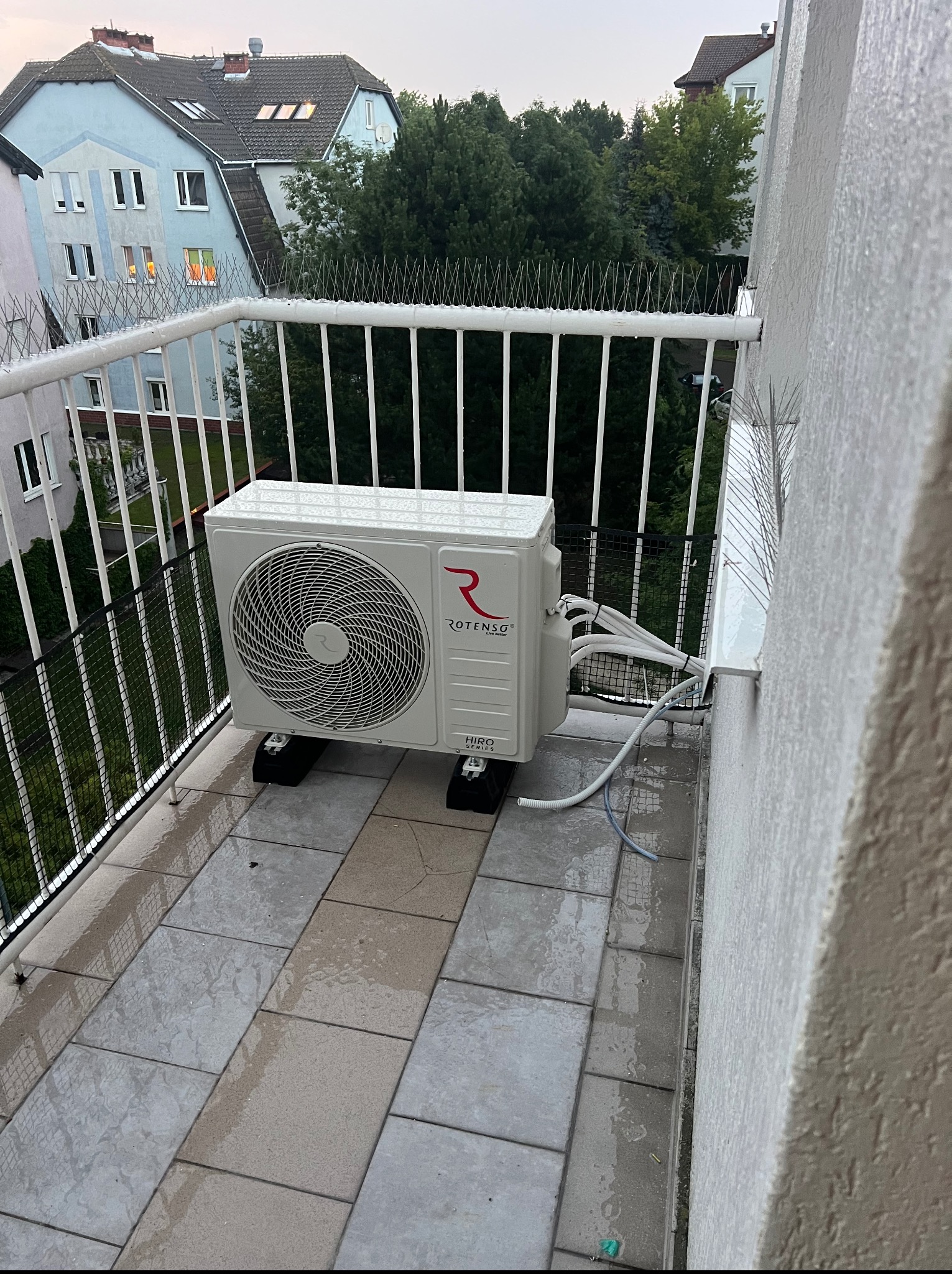 Biała jednostka zewnętrzna klimatyzacji Rotenso zamontowana na balkonie z szarymi płytkami, zabezpieczonym kolcami przeciw ptakom. Widok na budynki mieszkalne i zieleń.