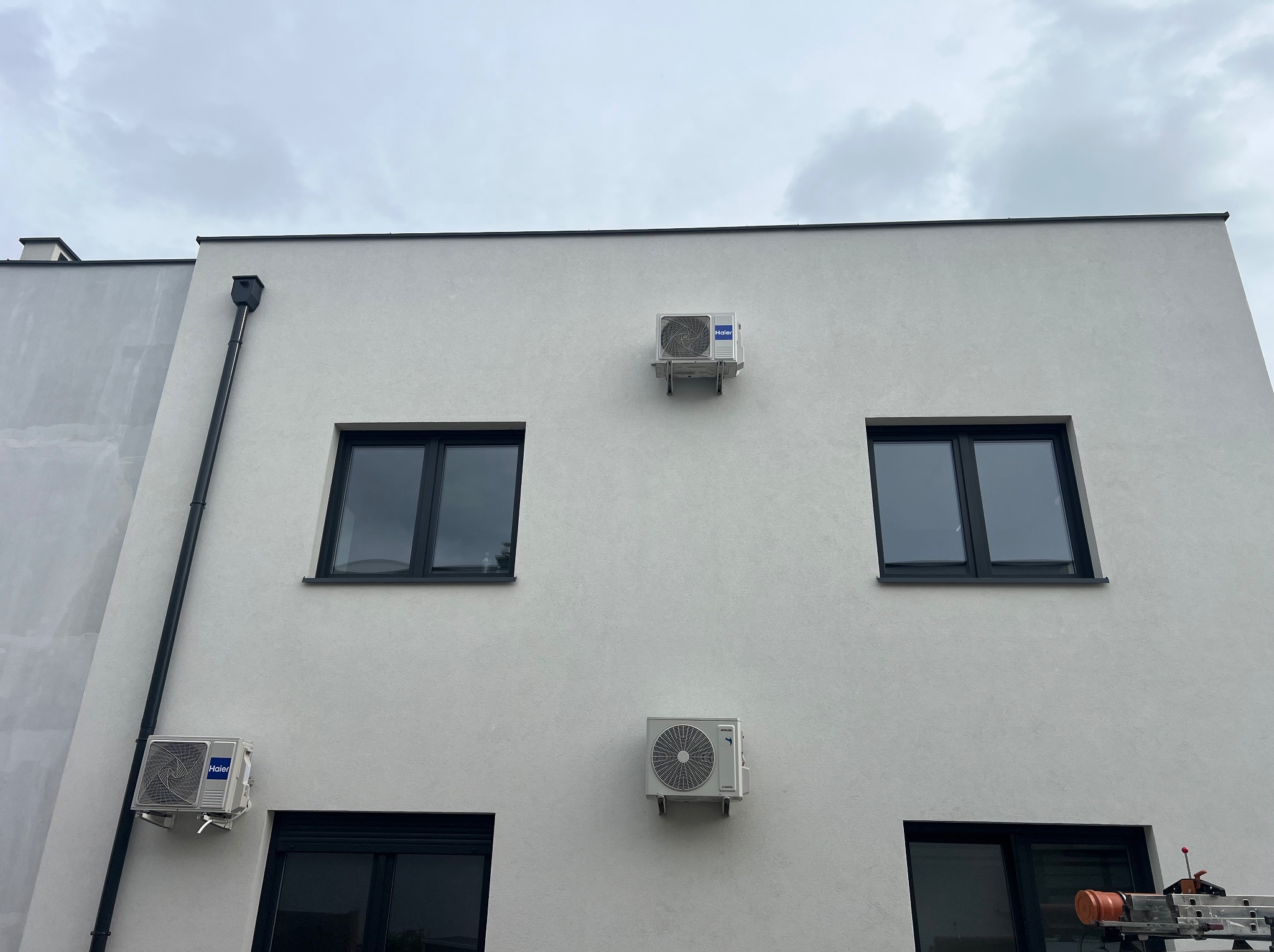 Elewacja budynku z trzema klimatyzatorami Haier, widoczne czarne okna, rynna i fragment drabiny. Pochmurne niebo w tle.
