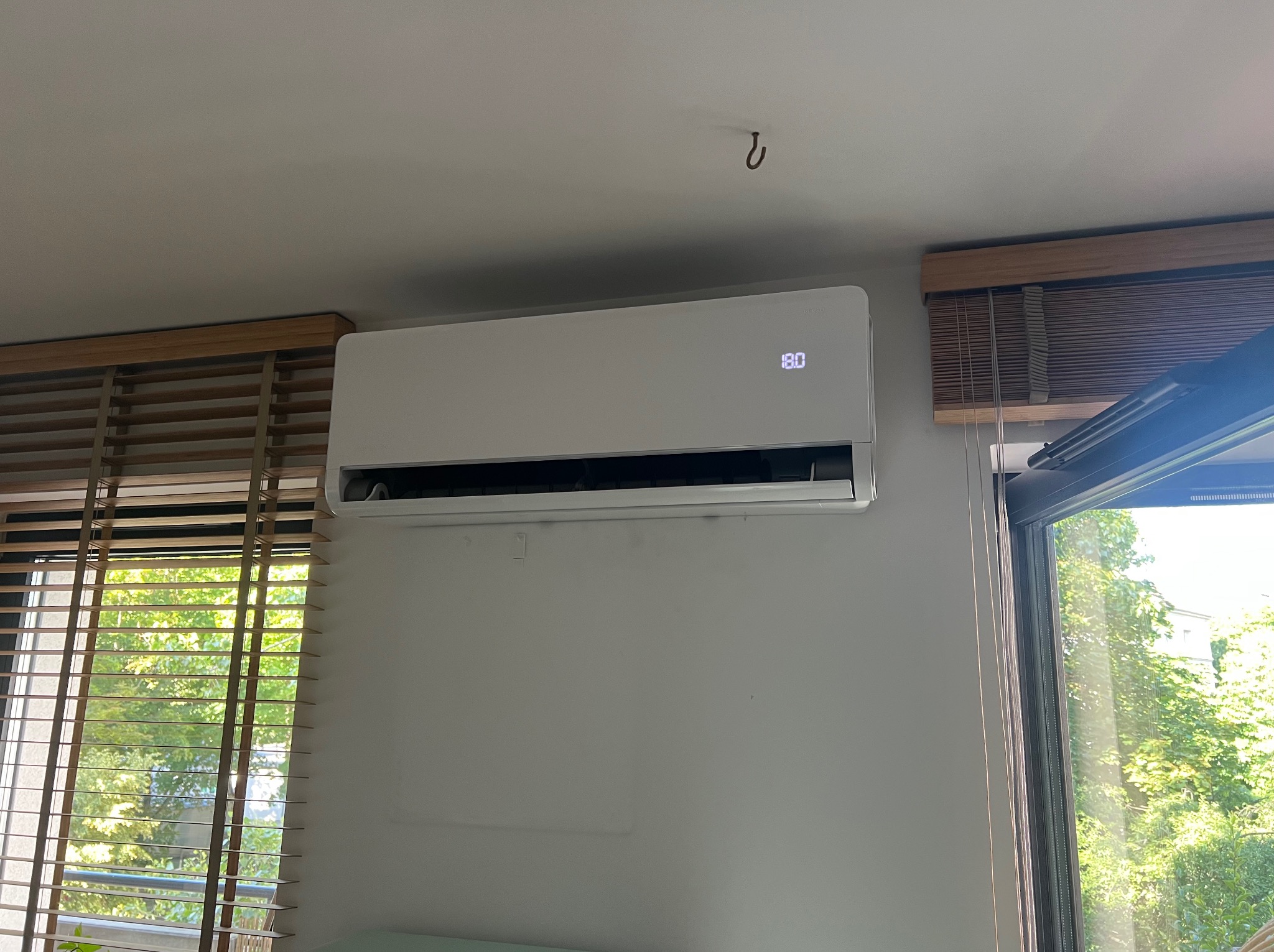 Biała klimatyzacja ścienna w pokoju z drewnianymi żaluzjami i widokiem na zieleń za oknem; wyświetlacz pokazuje 18 stopni Celsjusza.