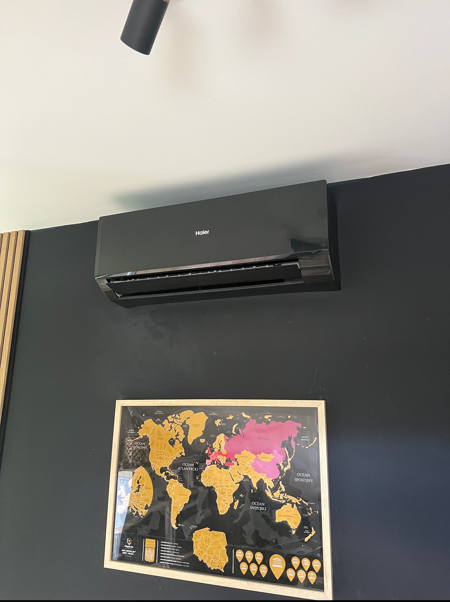 Czarna klimatyzacja Haier na ścianie nad mapą świata w Poznaniu. Nowoczesny design wnętrza z elementami drewna. Punktowe oświetlenie sufitowe.