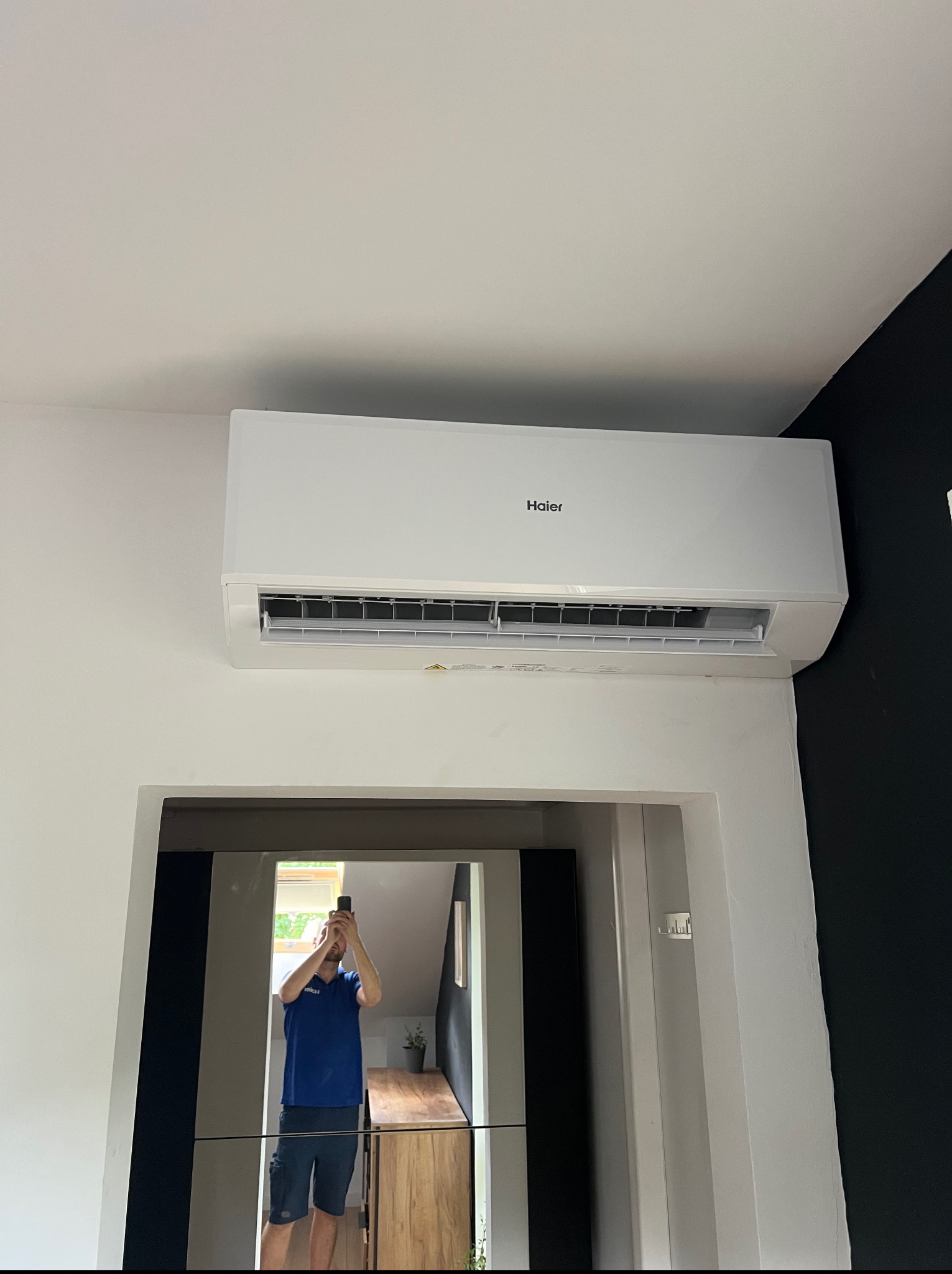 Biała klimatyzacja Haier, zamontowana na ścianie w jasnym wnętrzu. W lustrze odbija się osoba robiąca zdjęcie. Kontrastująca czarna ściana w tle.