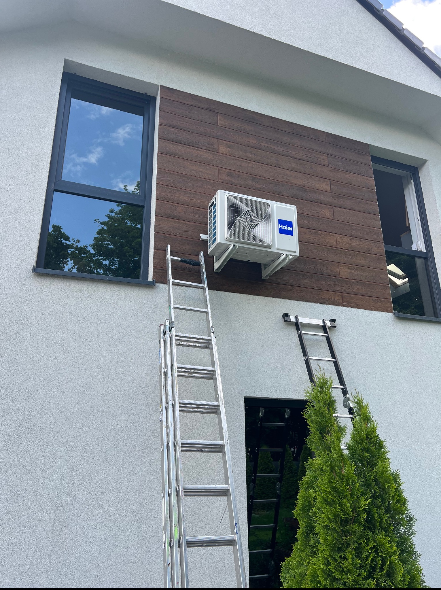 Biała klimatyzacja Haier zamontowana na elewacji domu z drewnianymi panelami, widoczna drabina, okna z ciemnymi ramami i fragment zielonego krzewu.