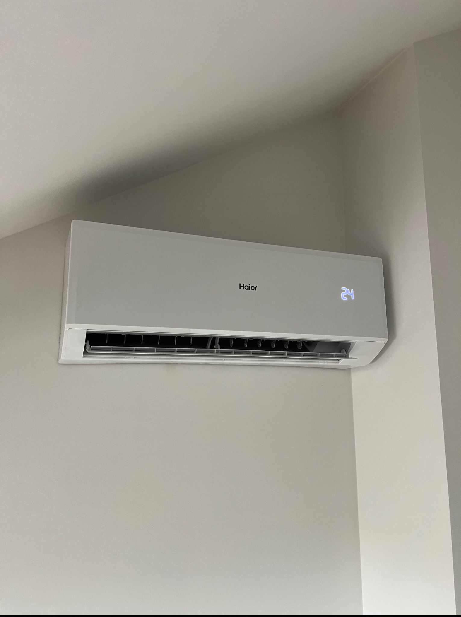 Biała klimatyzacja Haier na jasnej ścianie, wyświetlająca temperaturę 24 stopnie Celsjusza. Widok z dołu, oświetlenie naturalne.