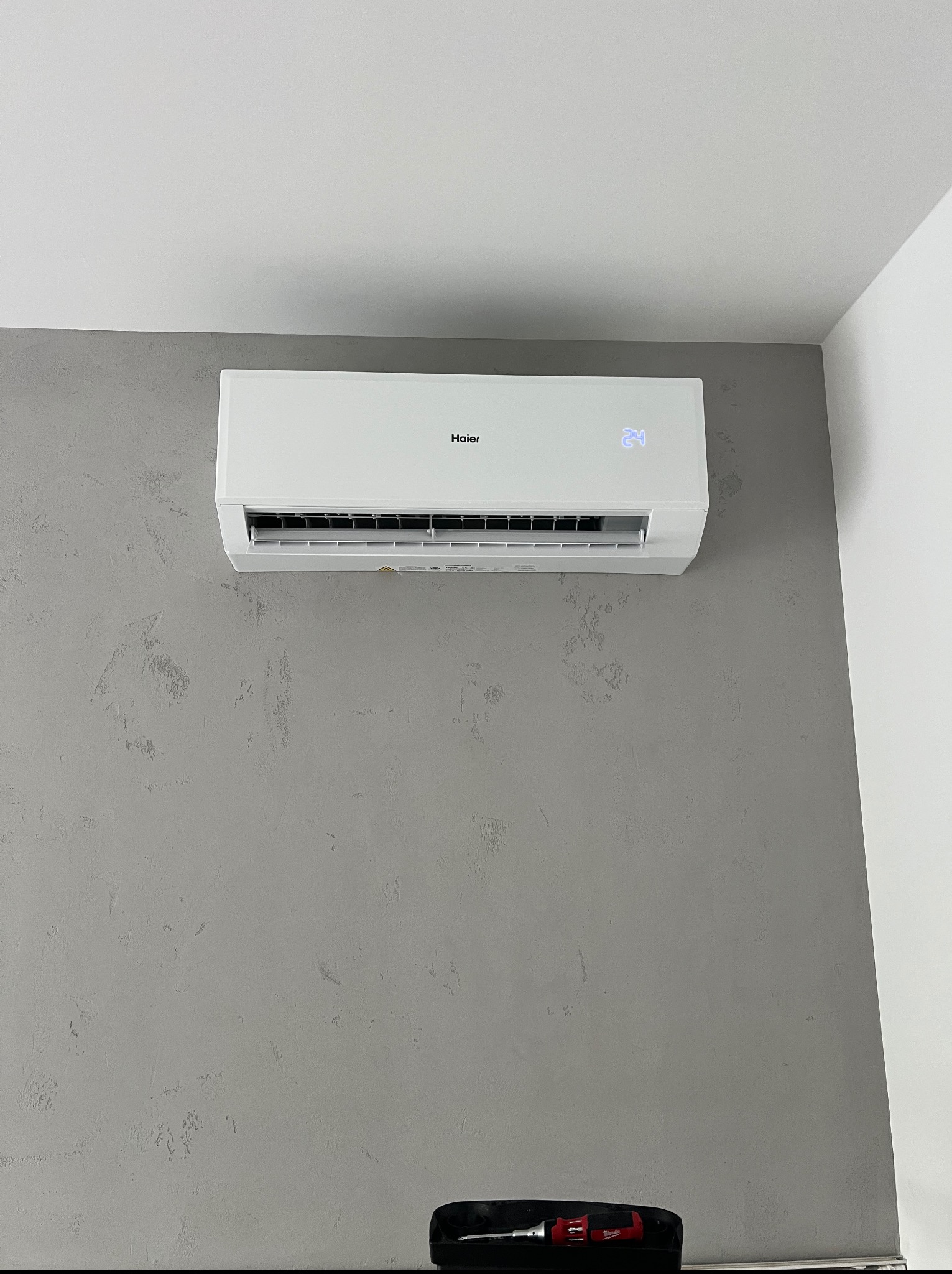 Biały klimatyzator Haier z wyświetlaczem temperatury 24°C, zamontowany na szarej ścianie o fakturze betonu. W tle widoczny fragment białej ściany i narzędzia.