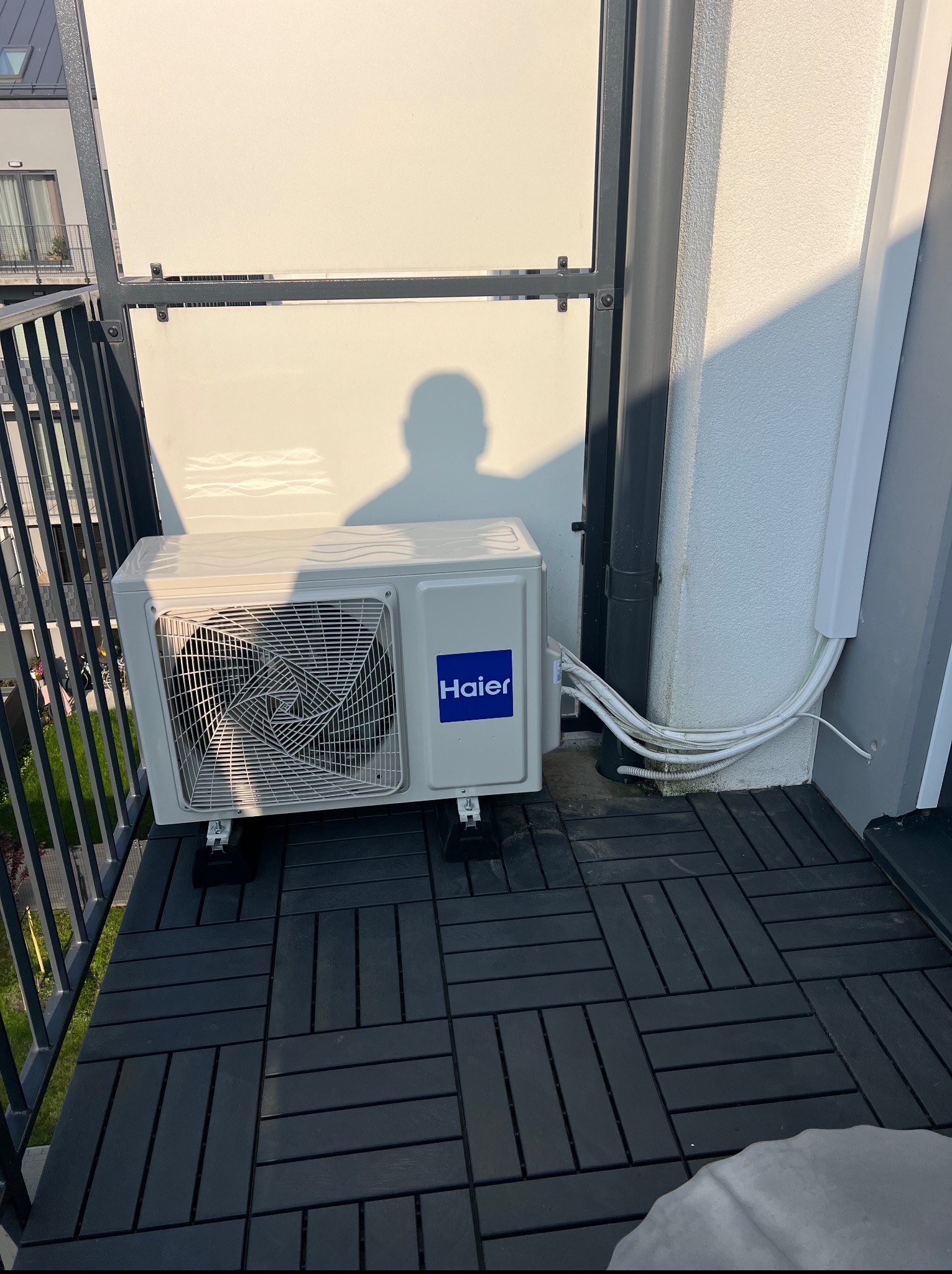 Biała jednostka zewnętrzna klimatyzacji Haier, zamontowana na balkonie z czarną podłogą, widok z boku, z widocznymi przewodami i cieniem osoby na ścianie.