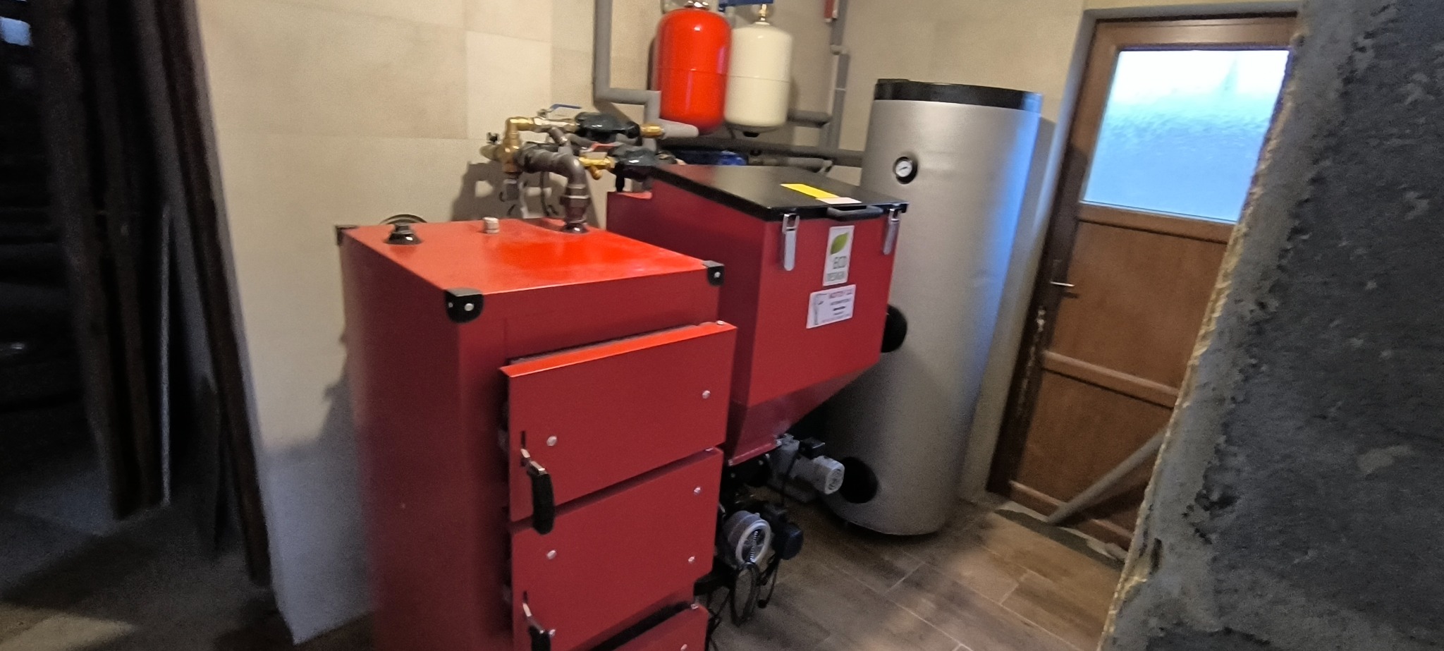 Czerwony piec na paliwo stałe z podajnikiem, zbiornik akumulacyjny i instalacja CO w kotłowni, widoczne rury i armatura.