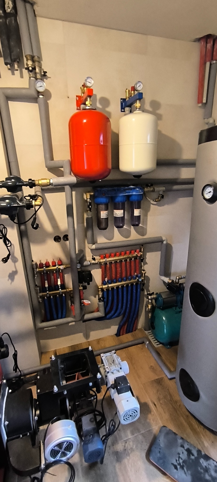Szara instalacja hydrauliczna i grzewcza z czerwonym i białym zbiornikiem wyrównawczym, filtrami wody, rozdzielaczami z rurami czerwonymi i niebieskimi oraz kotłem CO, zamontowana na ścianie...