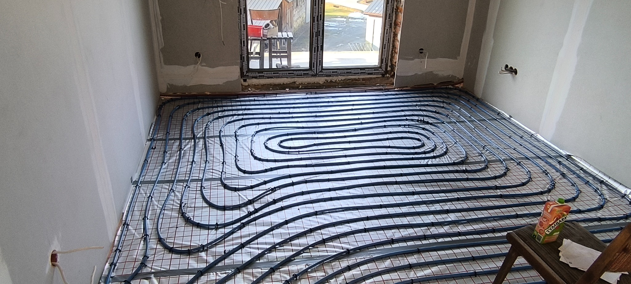 Niebieskie rurki ogrzewania podłogowego ułożone na siatce w surowym pomieszczeniu z oknem, karton soku na krześle w rogu.