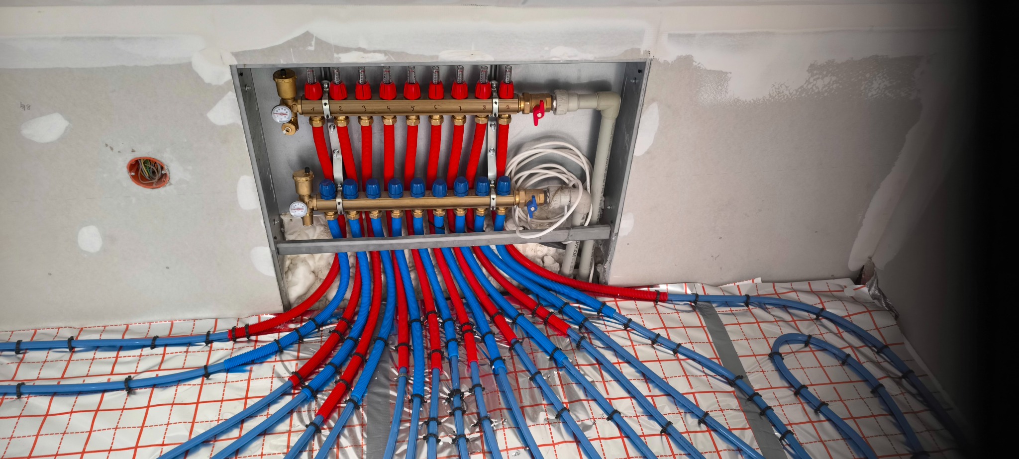 Rozdzielacz ogrzewania podłogowego z czerwonymi i niebieskimi rurami na folii izolacyjnej, widoczne manometry i instalacja w ścianie w trakcie montażu.