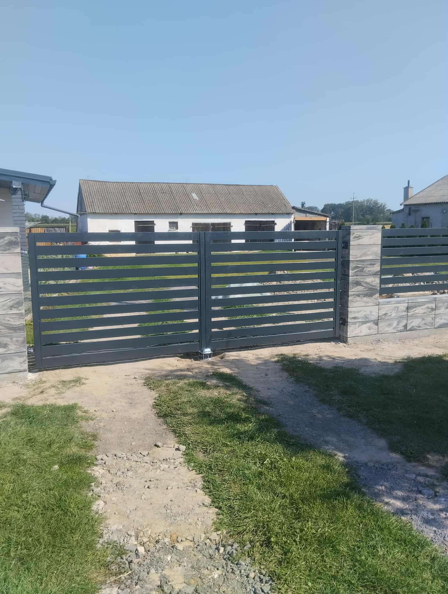 Nowoczesna, przesuwna brama w kolorze grafitowym z poziomymi panelami, zamontowana na tle jasnego budynku. Widoczne betonowe słupki i trawnik.