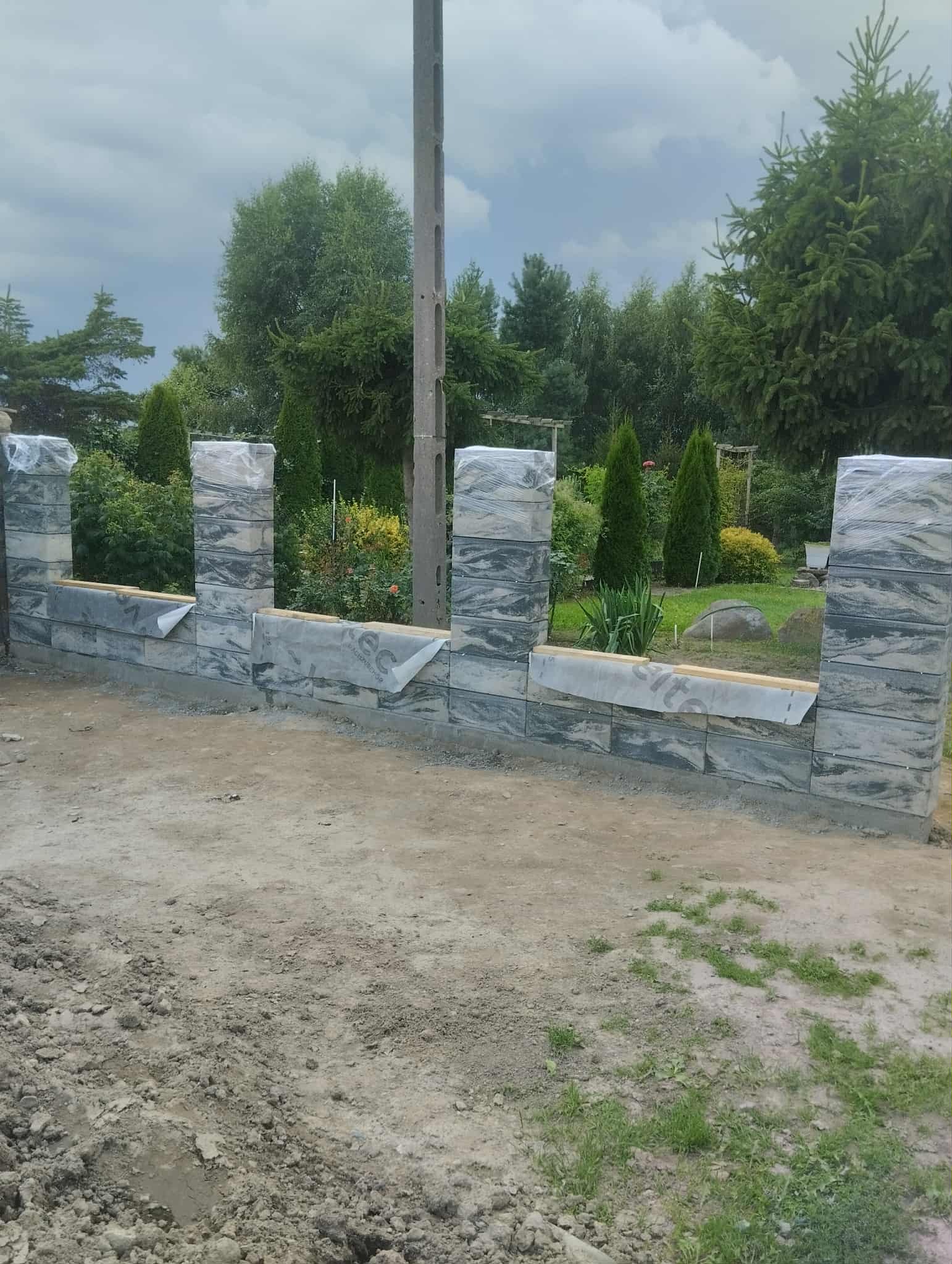 Wznoszony fragment ogrodzenia z szarej kostki łupanej, częściowo zabezpieczonej folią. Widoczne słupki i podmurówka. W tle zieleń ogrodu i słup energetyczny.