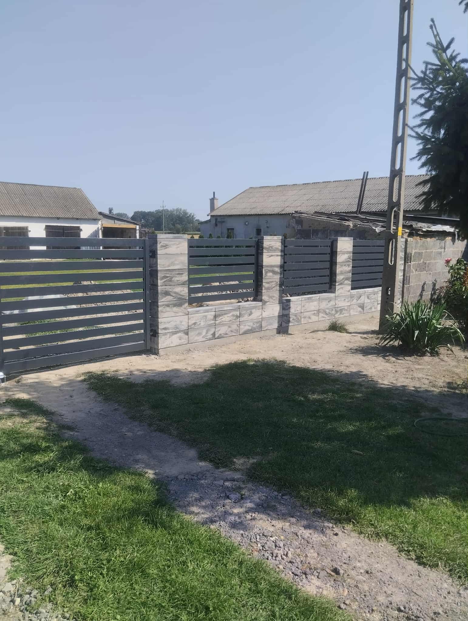 Nowoczesne ogrodzenie panelowe z szarymi przęsłami i słupkami z imitacji kamienia, na tle wiejskiego krajobrazu w słoneczny dzień. Widoczna brama wjazdowa.