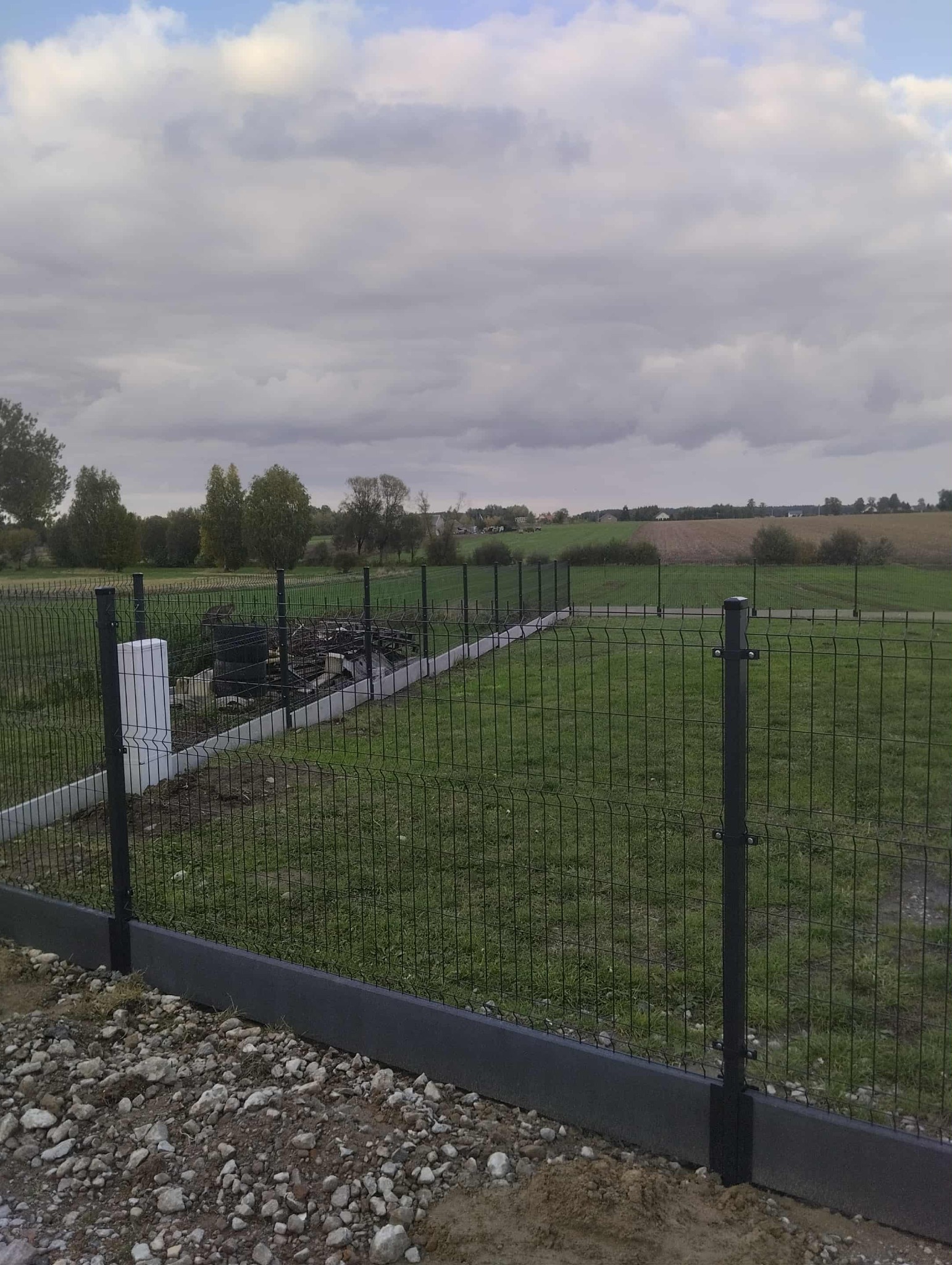 Nowoczesne, panelowe ogrodzenie w kolorze grafitowym z podmurówką, otoczone zielenią i polami uprawnymi w tle. Widoczny fragment terenu z gruzem.