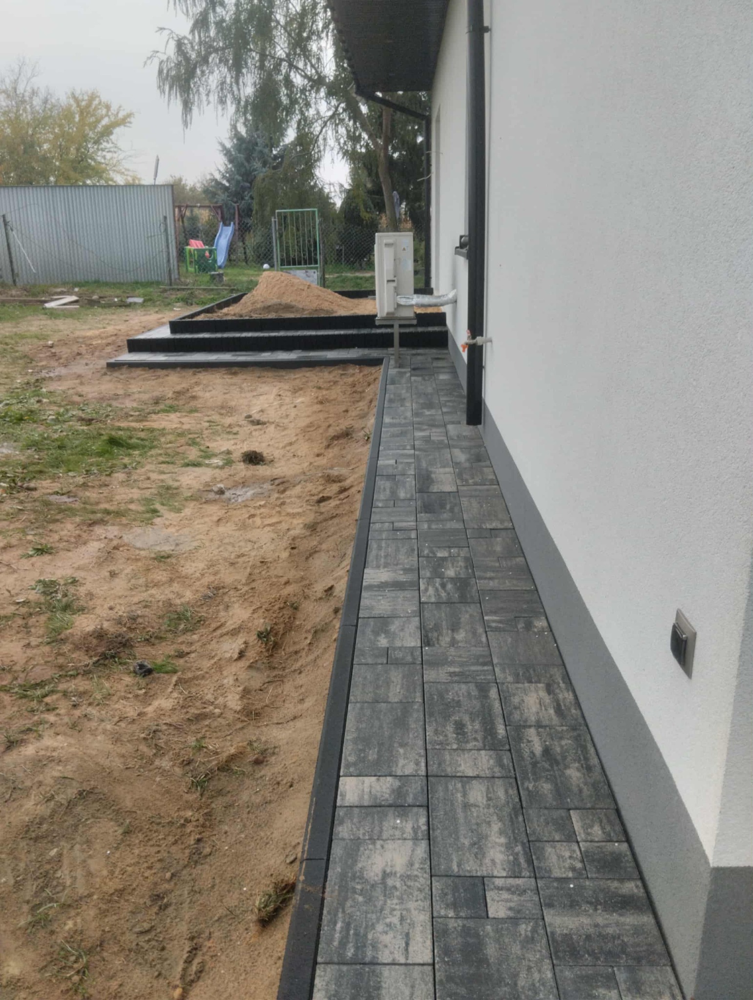 Ułożona kostka brukowa wzdłuż ściany domu, z widocznymi schodami z kostki i przygotowanym terenem pod dalsze prace. Szara kostka, biała ściana, ziemiste tło.