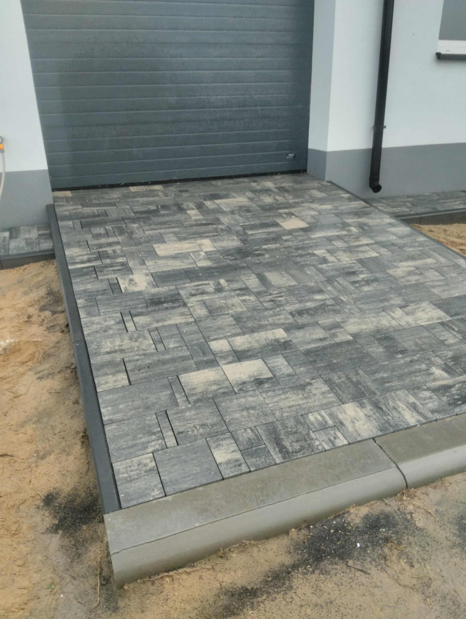 Ułożona kostka brukowa w odcieniach szarości prowadząca do garażu. Widoczne obrzeża i piasek. Kostka ułożona w nieregularny wzór, z bliska.