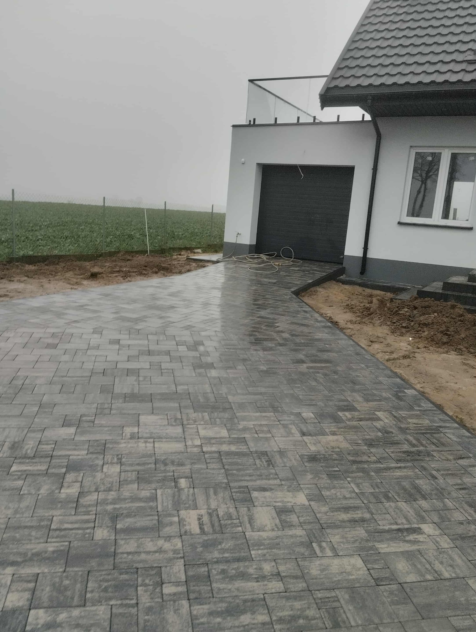 Ułożona, szara kostka brukowa prowadząca do nowoczesnego domu z garażem i szklaną balustradą balkonu. W tle widoczne pole i ogrodzenie w mglisty dzień.