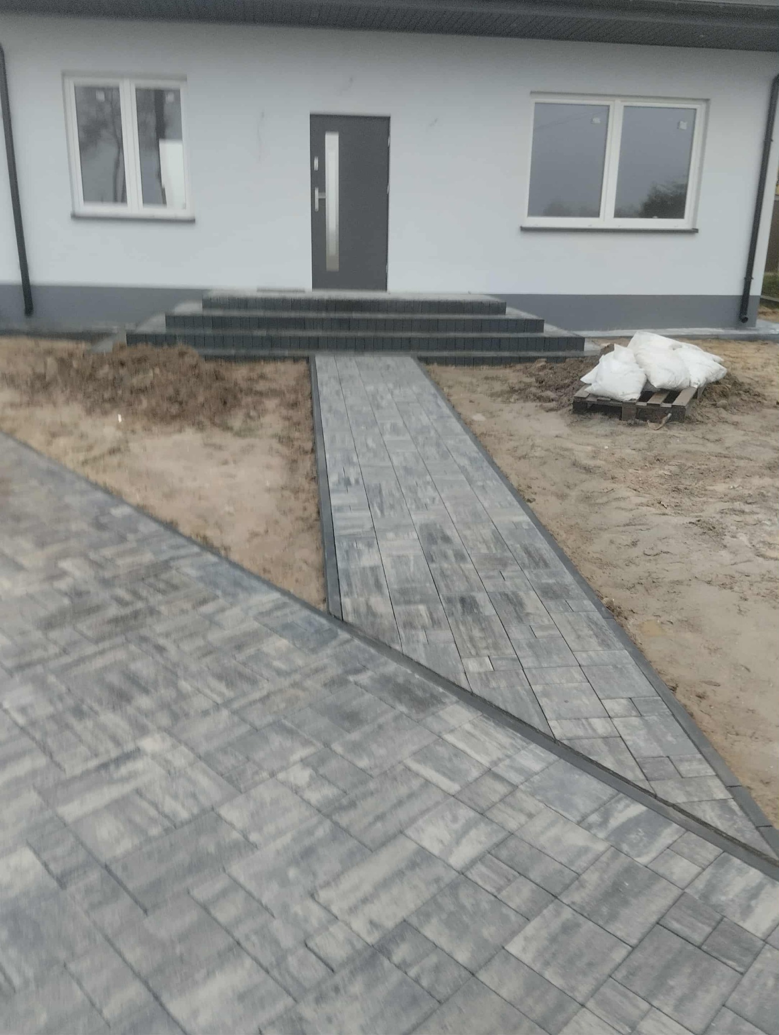 Nowo ułożona, szara kostka brukowa prowadząca do domu z szarymi drzwiami i schodami. Widoczne worki z materiałem budowlanym na palecie.