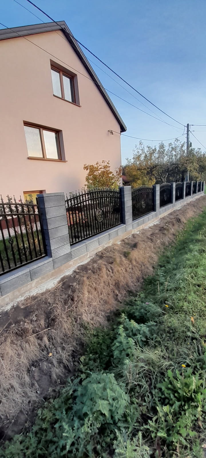 Eleganckie, czarne ogrodzenie z elementami dekoracyjnymi i szarymi słupkami z bloczków betonowych, oddzielające posesję od terenu zielonego. Widok na dom.