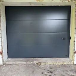 Nowa, antracytowa brama garażowa segmentowa w starej, odrapanej ramie z cegły i betonu. Widoczne trzy poziome segmenty i czarny uchwyt.