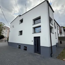 Odnowiona elewacja budynku z szarą podmur&oacute;wką i białymi ścianami. Widoczne okna z czarnymi ramami i ciemne drzwi wejściowe. Na dachu antena. Kostka brukowa na pierwszym planie.